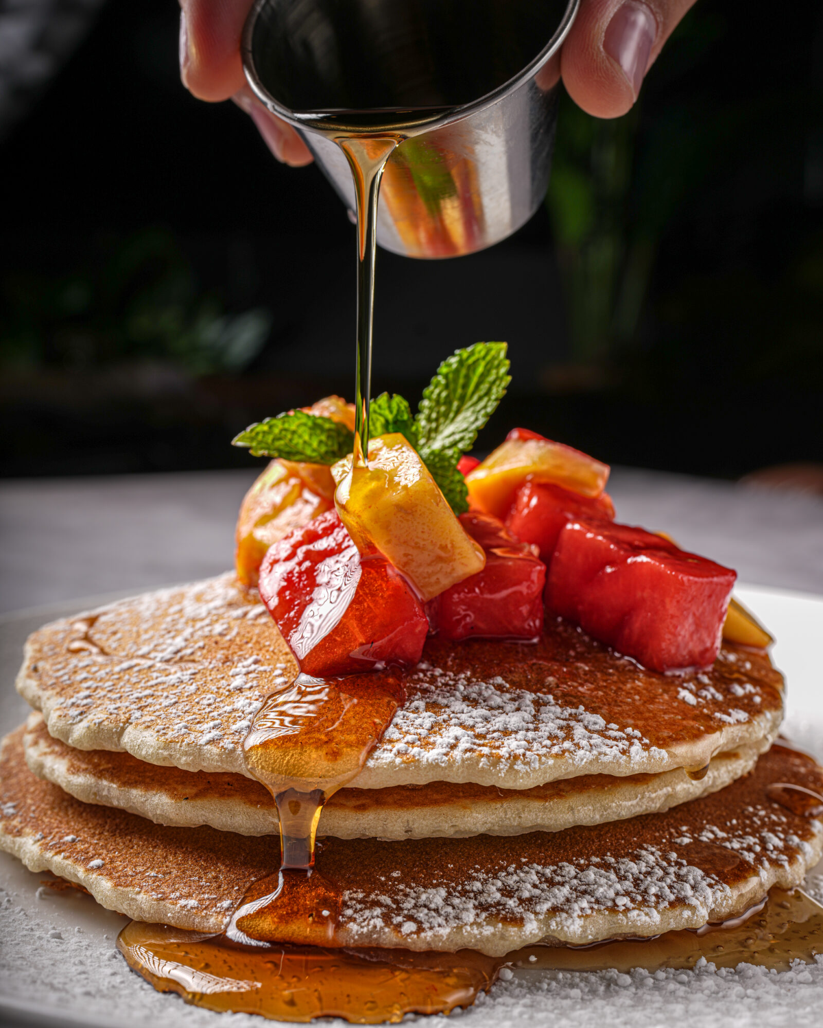 A tall stack of Fuego brunch pancakes topped with fresh fruit and a maple syrup pour